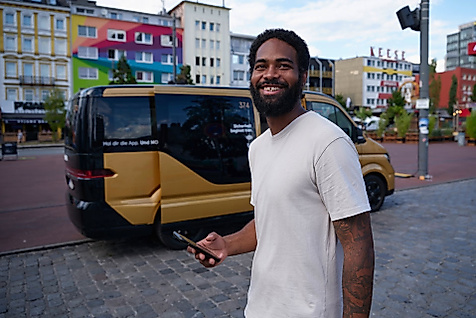 Mann mit Smartphone vor barrierefreiem goldfarbenem MOIA-Shuttle in urbaner Umgebung.