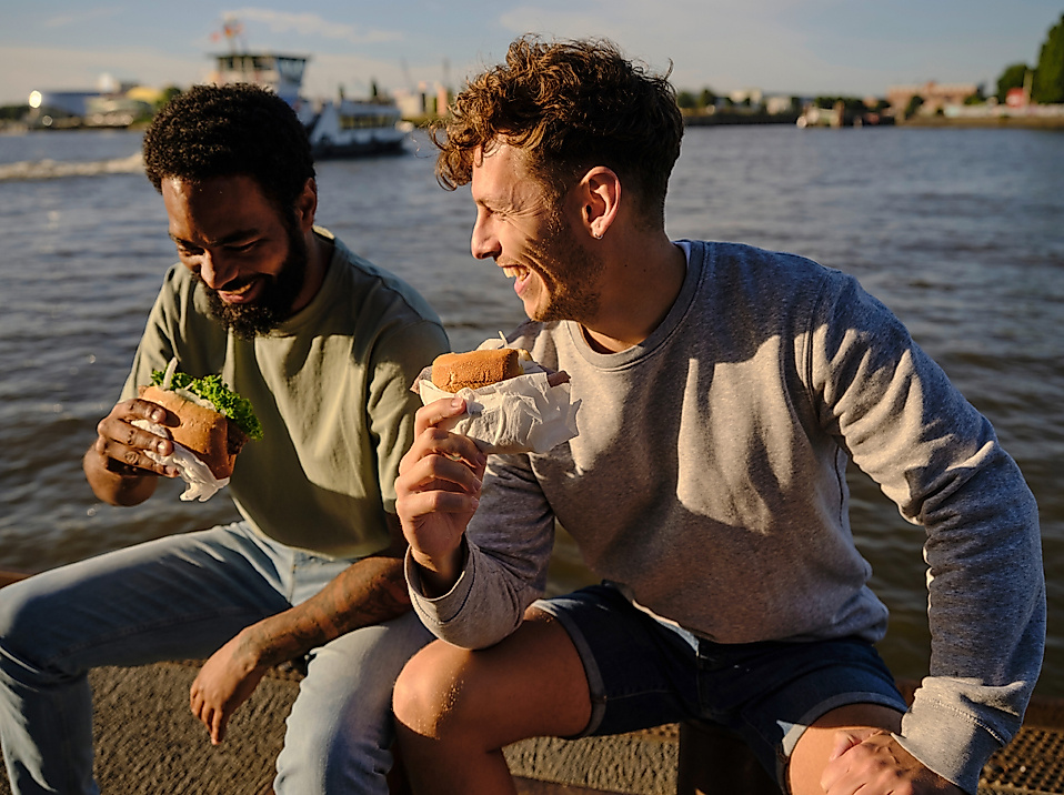 Zwei junge Männer sitzen entspannt am Wasserufer der Elbe in Hamburg und genießen Burger und Getränke im Sonnenlicht. Im Hintergrund ist ein Hafenboot auf dem Wasser zu sehen. Die Stimmung ist locker und sommerlich – ein typischer Genussmoment bei einem Picknick oder Snack mit Blick auf den Hafen, wie ihn Hamburg mit der Hamburg CARD erlebbar macht.