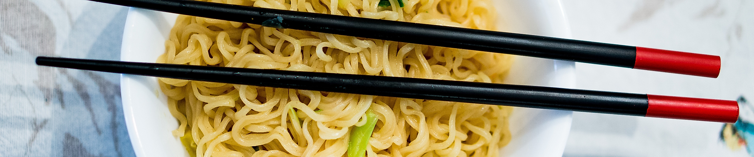 Schüssel mit asiatischen Nudeln, garniert mit Frühlingszwiebeln, dazu schwarze Essstäbchen mit roten Enden