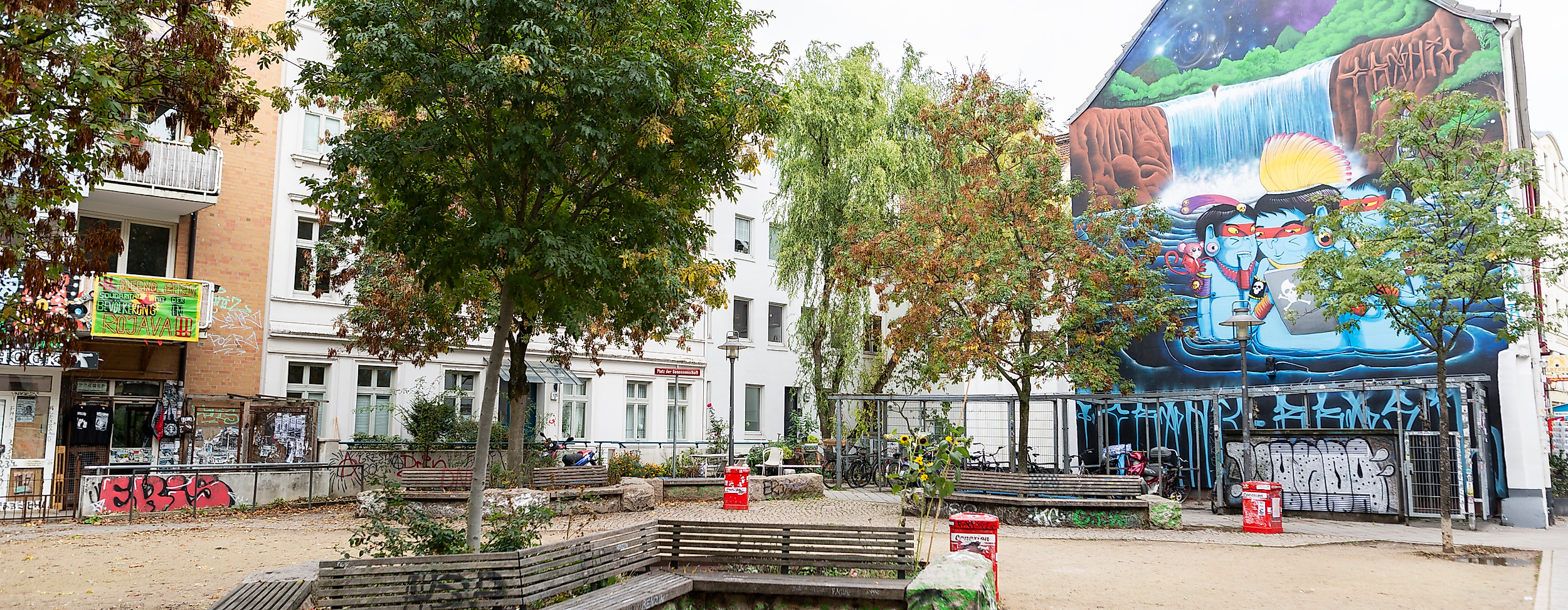 Platz im Hamburger Schanzenviertel mit Sitzbänken, Bäumen und farbenfrohem Wandgemälde an einer Giebelfassade