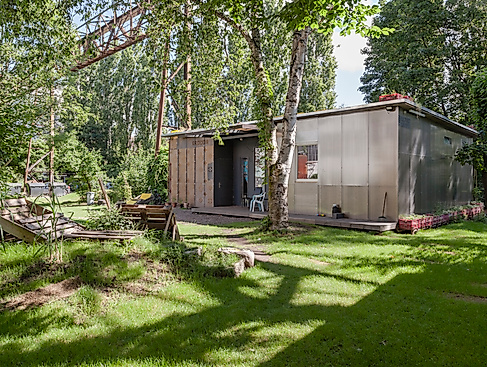 Photo of the Migrantpolitan at Kampnagel. A small one-storey house with a green garden, benches made of wooden pallets, surrounded by many trees.