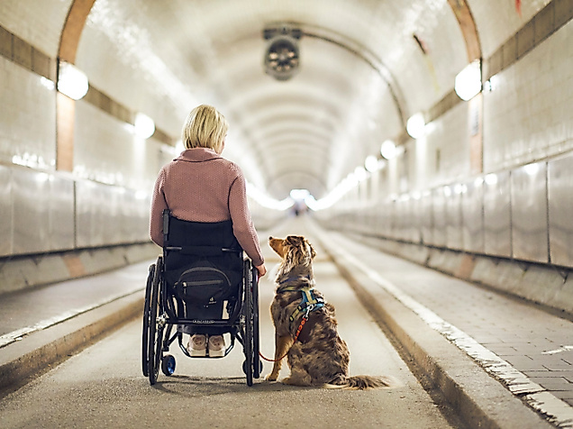 Rollstuhlnutzerin mit Assistenzhund im beleuchteten Alten Elbtunnel mit glatten Wegen und breitem Durchgang.