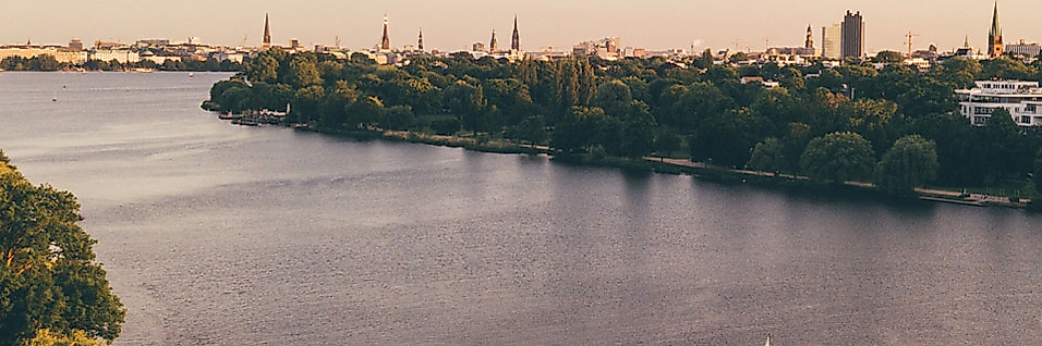 Segelboot auf der Außenalster bei Sonnenuntergang mit Blick auf Hamburgs grüne Ufer und Skyline.