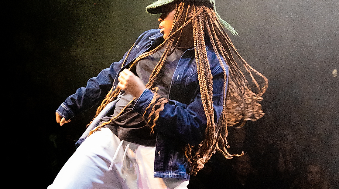 Eine Person mit Strickmütze, langen Braids und lockerer Kleidung steht auf einer Bühne und bewegt ihre Hüften mit gebeugten Knien in eine Richtung. Sie zieht sich mit dem gegenüberliegenden Arm in dieselbe Richtung hoch.