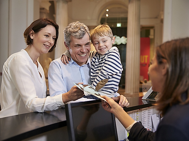 Frau, Mann und Kind, die an einem Tresen von einer Frau Infomaterial bekommen