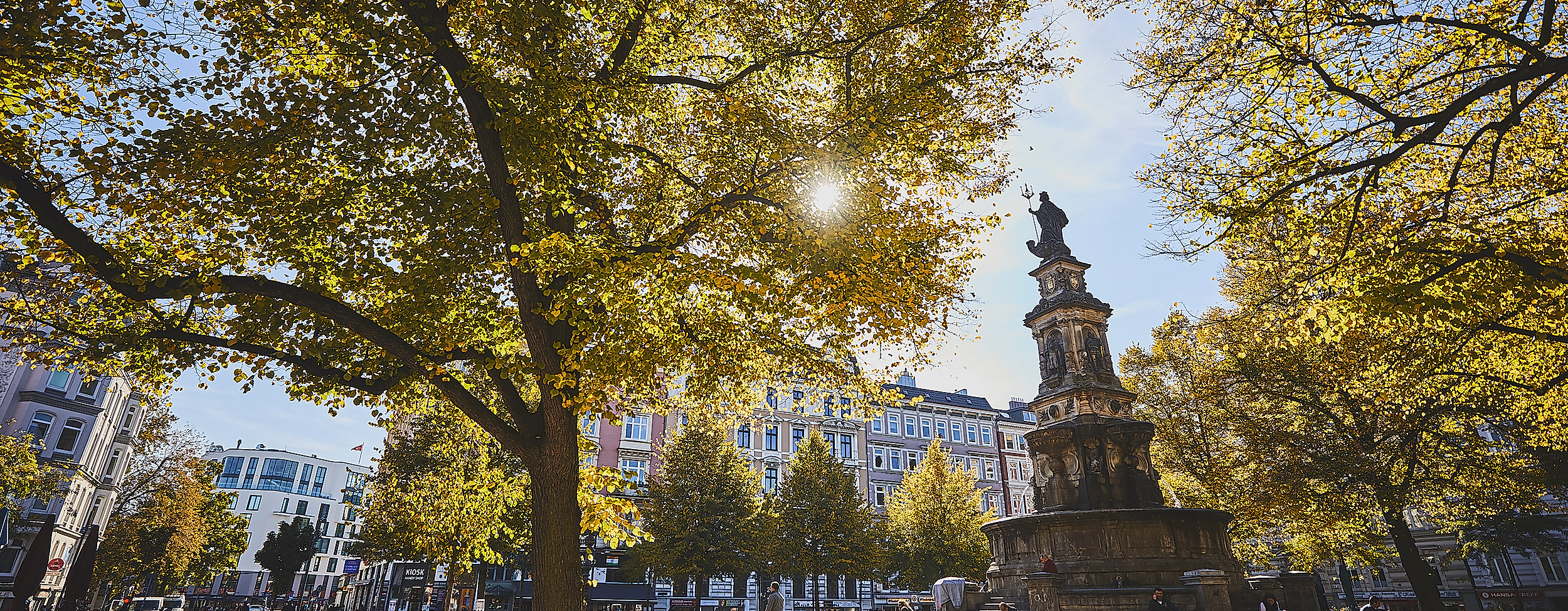 Hansaplatz in Hamburg-St. Georg mit St.-Georg-Brunnen, herbstlich gefärbten Bäumen und Sonnenlicht zwischen dem Laub