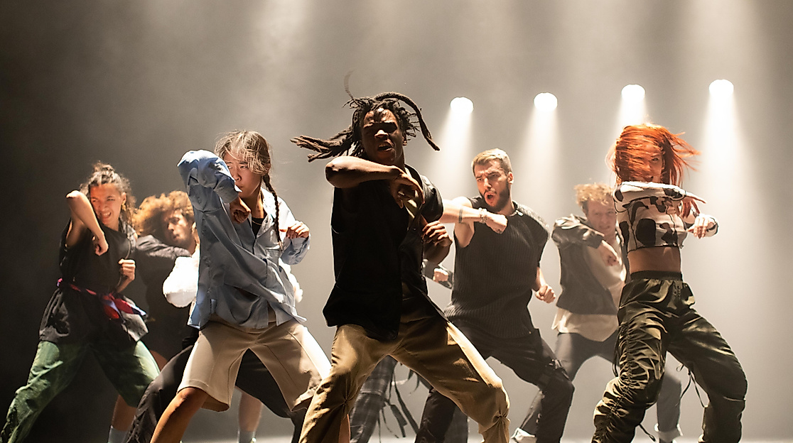 An energetic dance group performs a powerful choreography on stage under dramatic lighting.