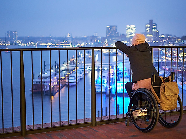 Rollstuhlnutzerin blickt abends von barrierefreier Plattform auf die beleuchtete Elbphilharmonie und den Hafen.