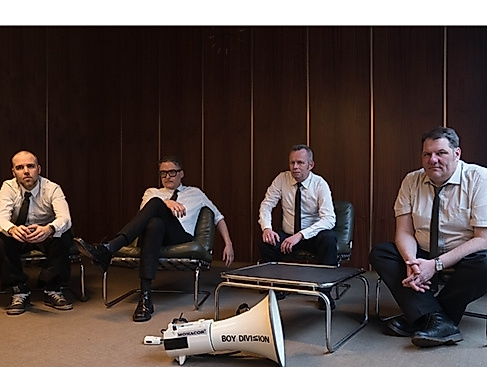 The four members of Boy Division all wear a white shirt and a narrow black tie; they sit casually in a '60s-style conference room, with a white megaphone in front of them, labeled with the band's name.
