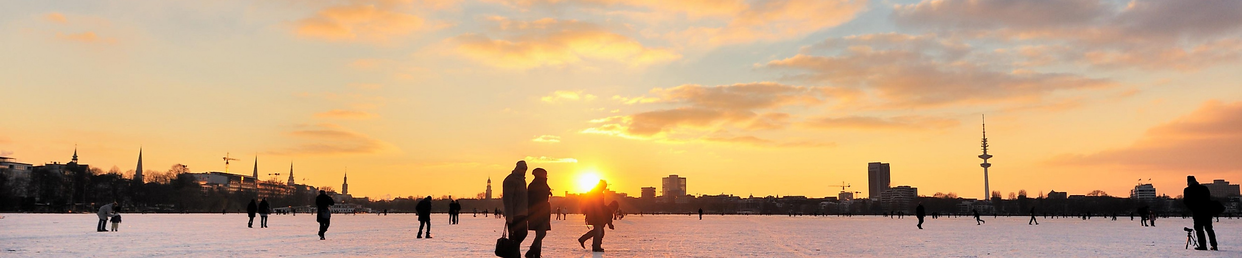 Menschen laufen Schlittschuh auf zugefrorener Alster bei Sonnenuntergang mit Blick auf Hamburgs Skyline