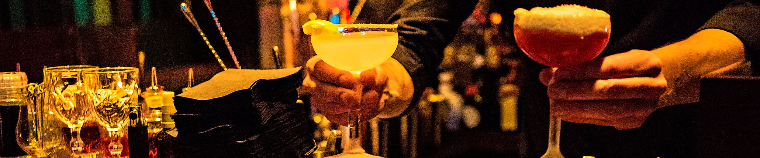 Zwei Cocktails werden an einer Bar bei warmem Licht serviert, daneben Gläser und Barutensilien