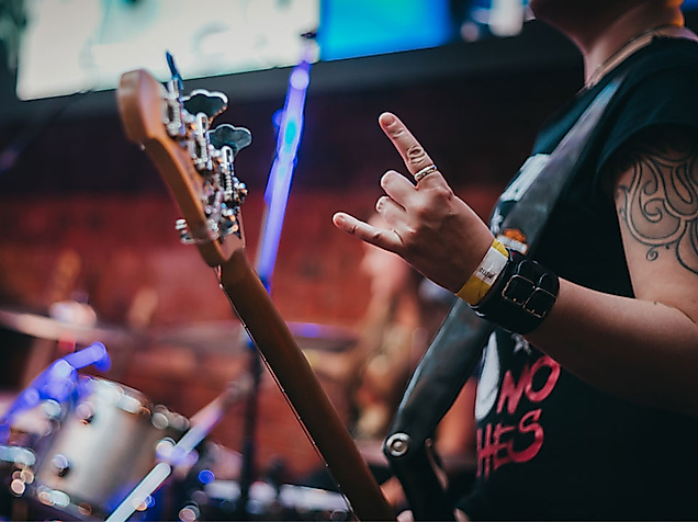 Gitarrist mit tätowiertem Arm und Rockhand-Geste auf Bühne, im Hintergrund ein Schlagzeug