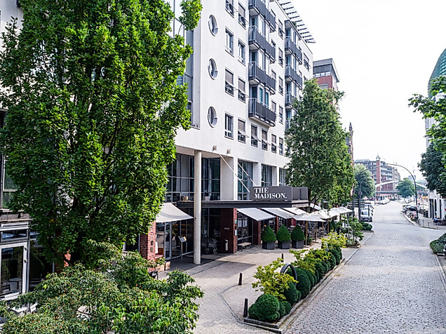 Moderne Häuserreihe mit Außengastronomie an gepflasterter Straße, umgeben von Bäumen.