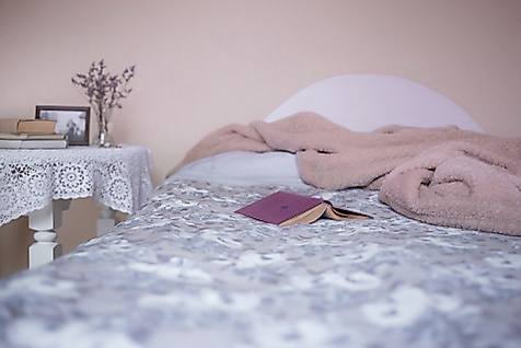 Offenes Buch liegt auf gemachtem Bett mit rosa Decke, daneben Nachttisch mit Büchern und Trockenblumen.