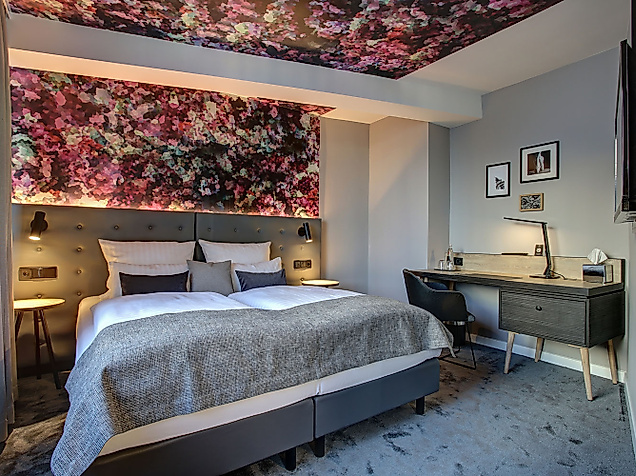 Modernes Hotelzimmer mit Doppelbett, floralem Muster an Wand und Decke sowie Schreibtisch mit Lampe.
