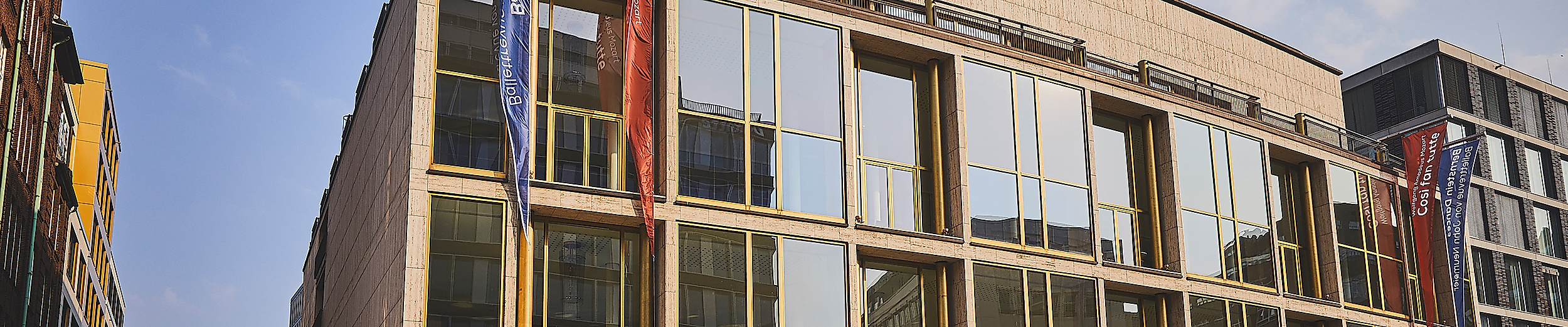 Moderne Glasfassade der Hamburgischen Staatsoper bei Tag mit Fahnen und Autos im Vordergrund.