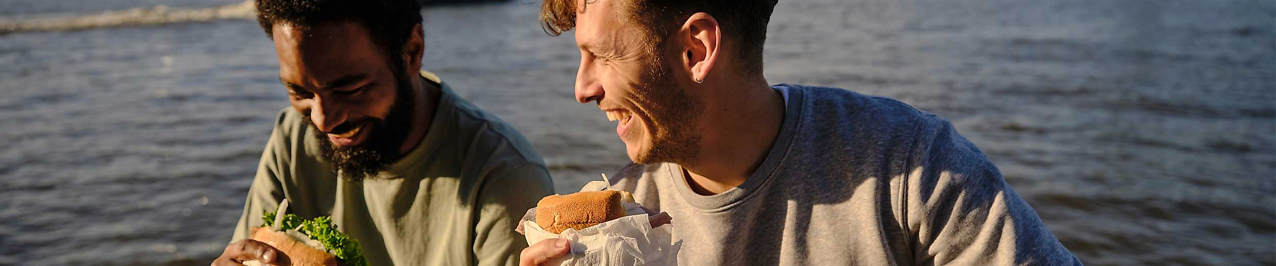 Zwei junge Männer sitzen entspannt am Wasserufer der Elbe in Hamburg und genießen Burger und Getränke im Sonnenlicht. Im Hintergrund ist ein Hafenboot auf dem Wasser zu sehen. Die Stimmung ist locker und sommerlich – ein typischer Genussmoment bei einem Picknick oder Snack mit Blick auf den Hafen, wie ihn Hamburg mit der Hamburg CARD erlebbar macht.