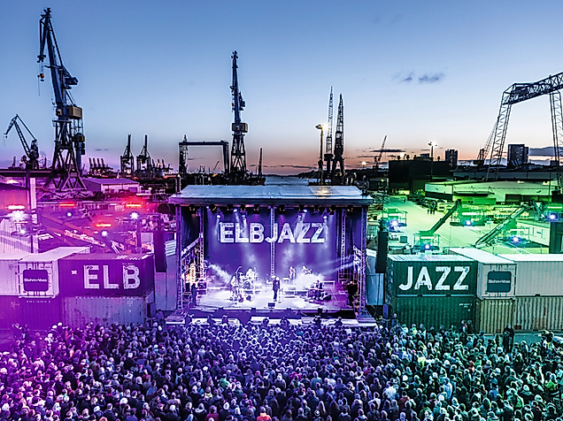 Beleuchtete Bühne des ELBJAZZ Festivals im Hamburger Hafen mit Menschenmenge und Hafenkränen bei Abenddämmerung.