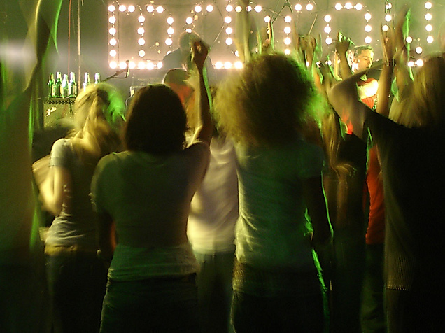 Feiernde Menschen tanzen im grünen Licht eines Elektroclubs mit leuchtender „Dance“-Schrift im Hintergrund