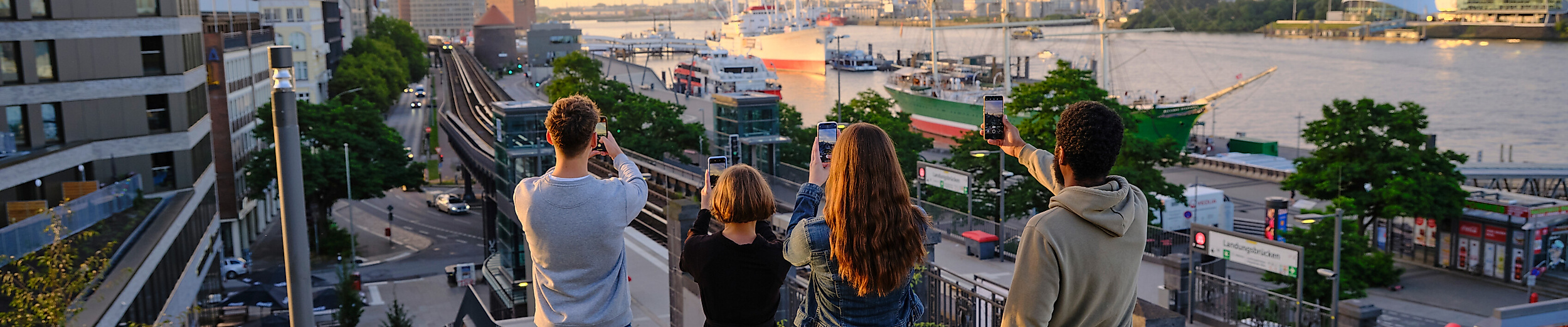 A group of young adults are taking photographs of the Hamburg skyline with their smartphones.