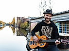 Mann mit Gitarre und Hut sitzt am Wasser vor Hausboot in Hamburg, lächelt in die Kamera bei Herbstlicht