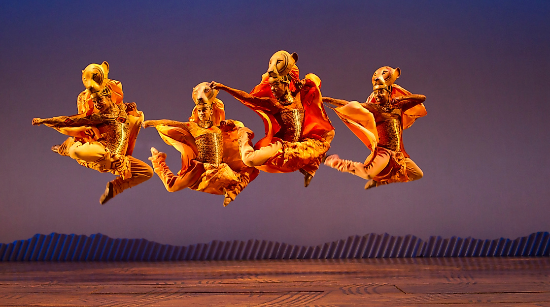 Szenenbild König der Löwen: Vier Tänzer:innen in Löwenkostümen springen synchron vor afrikanischer Savannenkulisse.